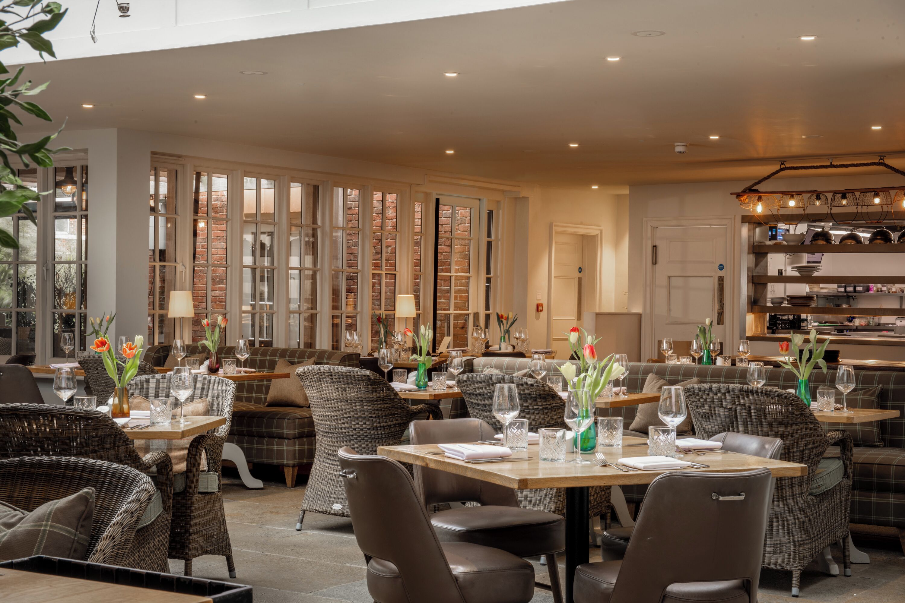 A spacious indoor dining area with wicker furniture and tables set for dining, featuring a brick wall and large windows.