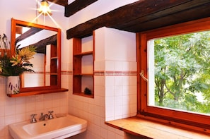 Un baño con techo de vigas de madera, un lavamanos blanco, un espejo y una ventana con vistas a árboles.