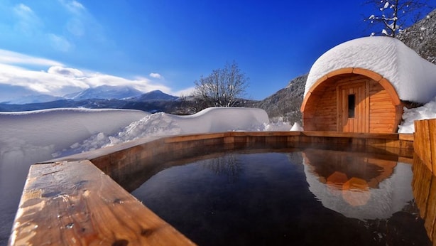 Un bain à remous en bois, dont le toit est recouvert de neige, entouré d’un paysage enneigé et de montagnes.