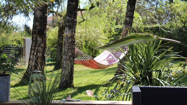 Eine Hängematze, zwischen zwei Bäumen in einem Garten mit Blumentöpfen und einer Holzbank befestigt.