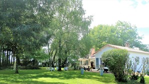 Ein Haus mit weißer Fassade, einem roten Ziegeldach und einer kleinen Veranda. Mehrere Bäume befinden sich im Garten, und einige Stühle sind zu sehen.