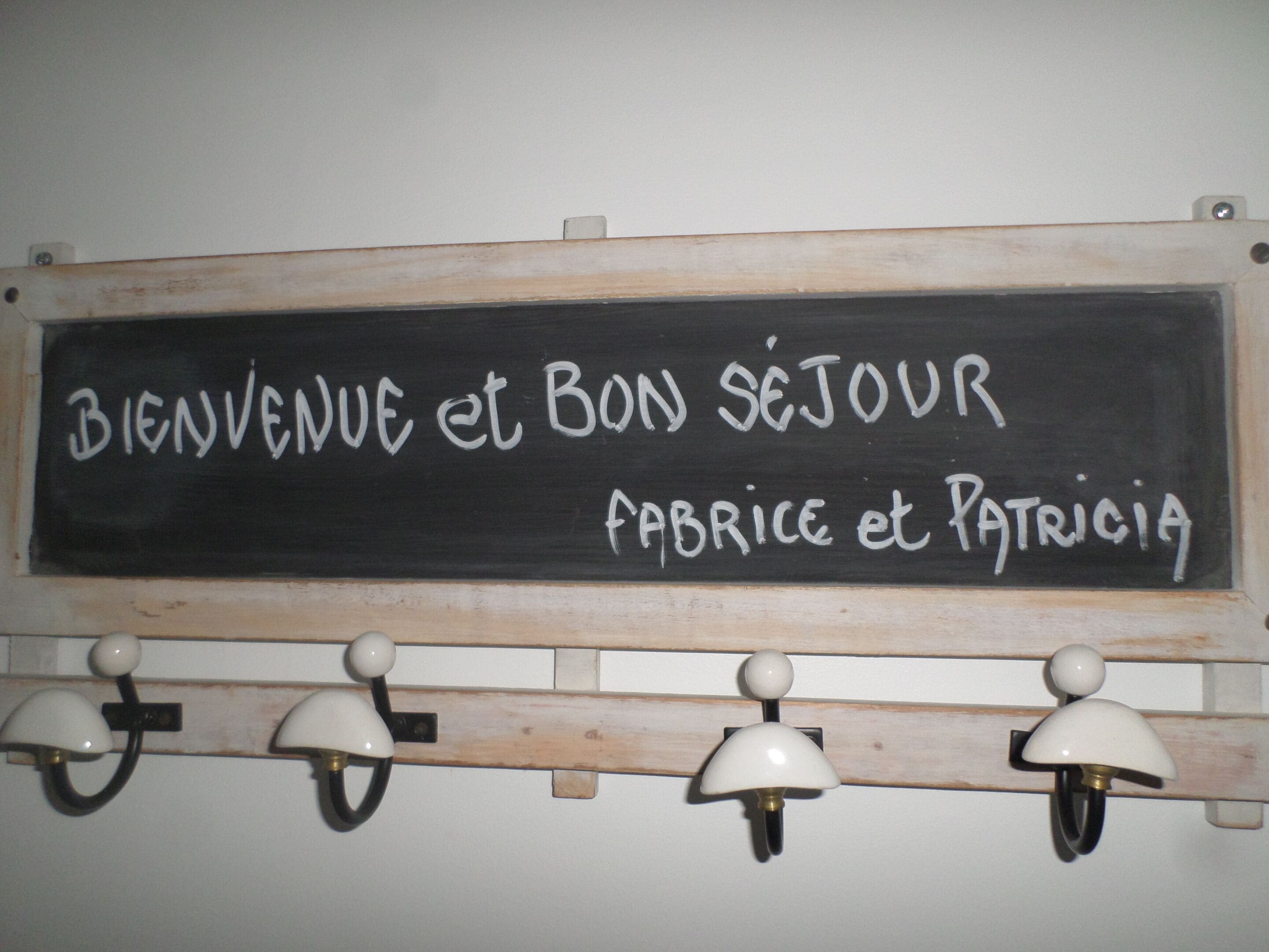 Un tableau noir sur lequel on peut lire « Bienvenue et bon séjour » et « Fabrice et Patricia », fixé au mur avec trois lampes murales en dessous.