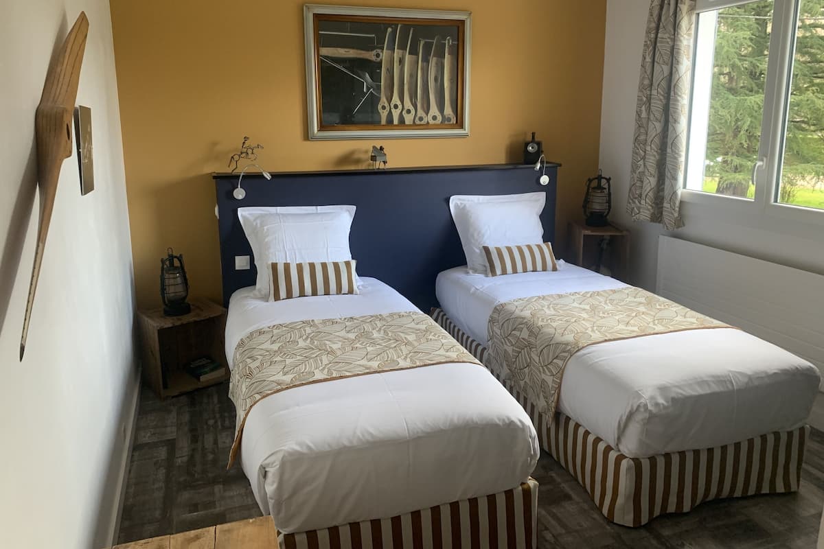 Habitación doble con dos camas, un cuadro enmarcado sobre la cabecera, una ventana con cortinas y una mesita con un farol.