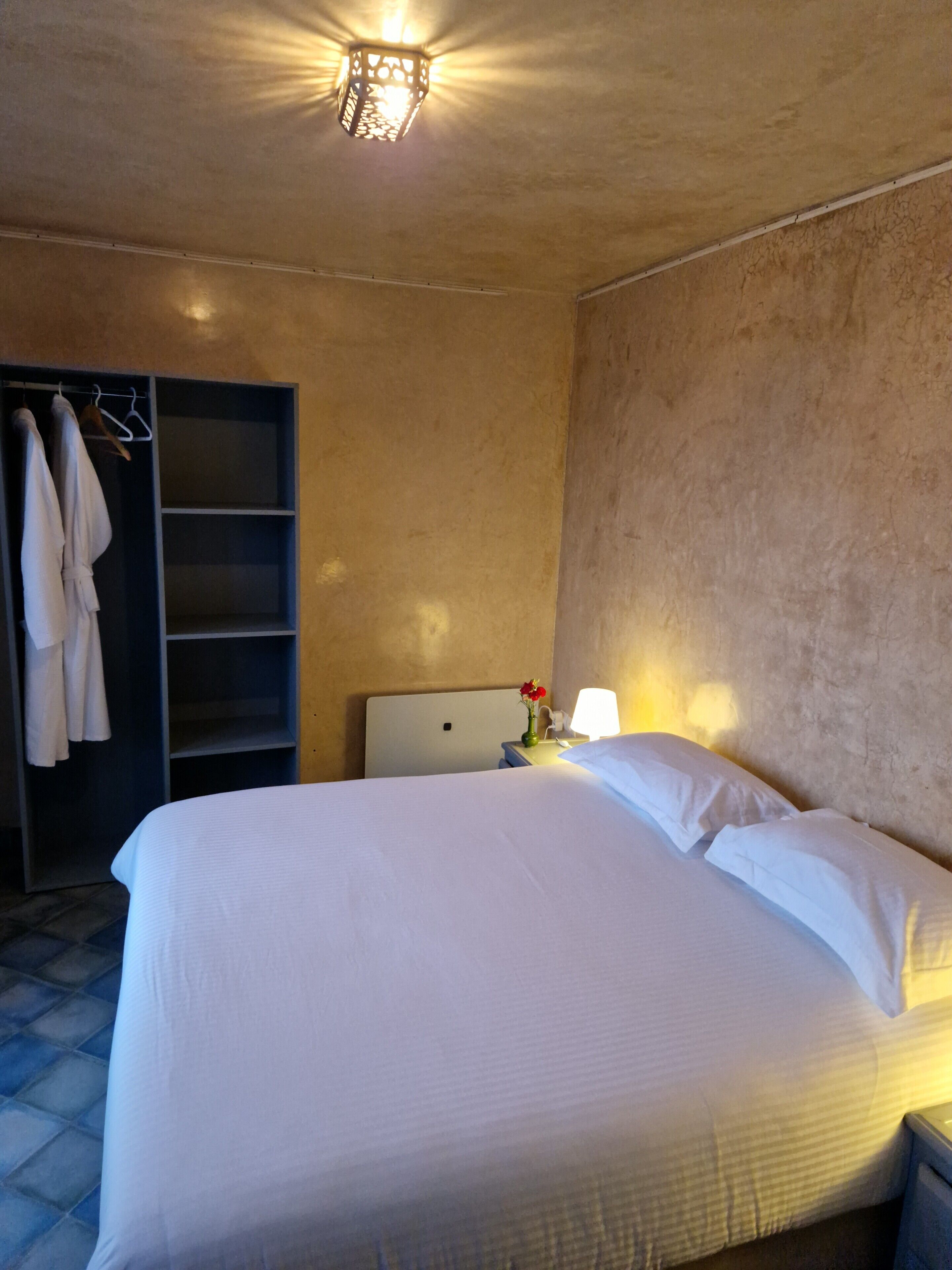 A hotel room with a large bed, a wardrobe with hanging clothes, a nightstand with a lamp, and a ceiling light fixture.
