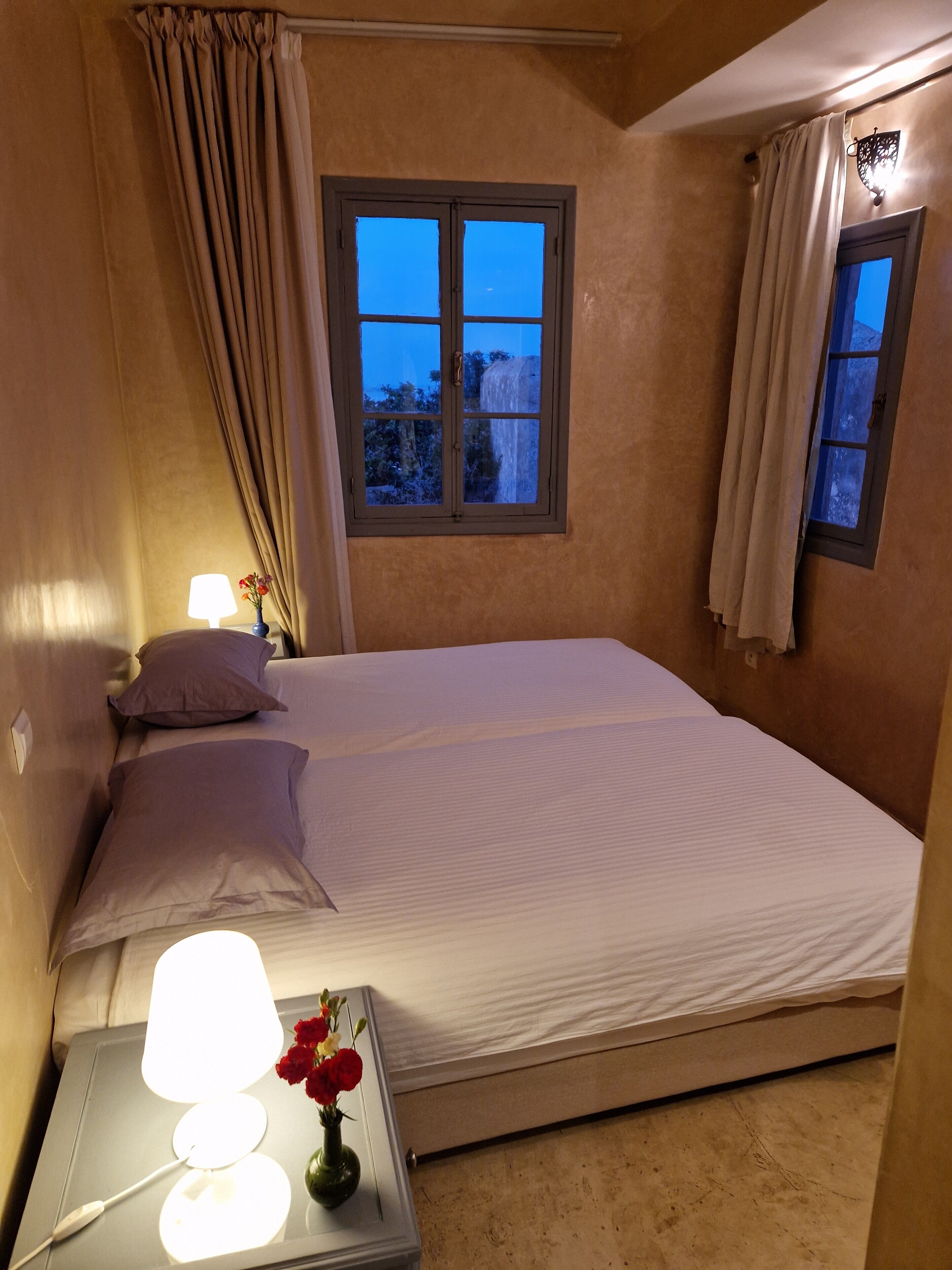 Une chambre à coucher avec un lit, deux lampes de chevet, une table de chevet ornée d’un vase de fleurs et une fenêtre avec des rideaux.