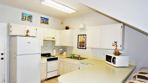 A compact kitchen with white cabinets, a refrigerator, a microwave, and a sink. There are decorative items on the countertops and a small island with chairs. - 108 W. Morningside 2 Bedroom Condo by RedAwning (South Padre Island)