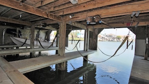 A wooden pier with a boat docked on the left, a hammock hanging from the ceiling, and a calm water body in the background. - 108 W. Morningside 2 Bedroom Condo by RedAwning (South Padre Island)