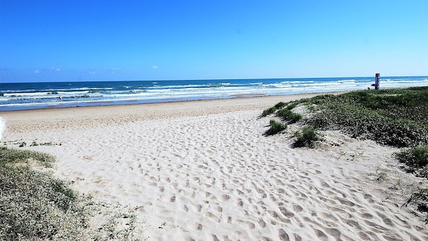 A sandy beach with footprints leading to the water, a clear sky, and a structure in the distance. - 108 W. Morningside 2 Bedroom Condo by RedAwning (South Padre Island)