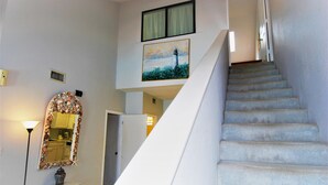A staircase with a painting of a lighthouse above it, a mirror with a seashell frame, and a small table with a vase. - 108 W. Morningside 2 Bedroom Condo by RedAwning (South Padre Island)