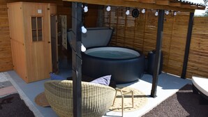 Un espace extérieur aménagé avec un bain à remous en bois, surmonté d’une pergola, comprenant une chaise en osier et un meuble en bois.