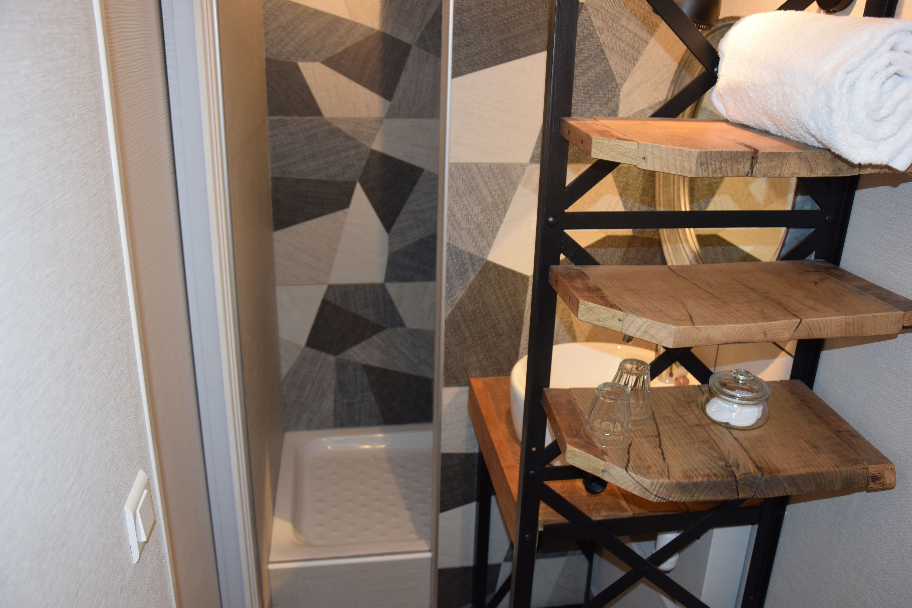 Une salle de bain avec un mur de douche orné d’un motif géométrique, une étagère en bois sur laquelle est posée une serviette roulée, et un petit bocal en verre.