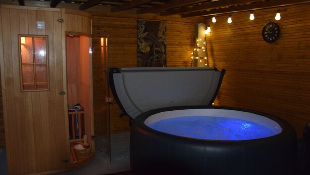 Une cabine de sauna en bois, avec une grande baignoire noire remplie d’eau, une porte en bois et une horloge murale.