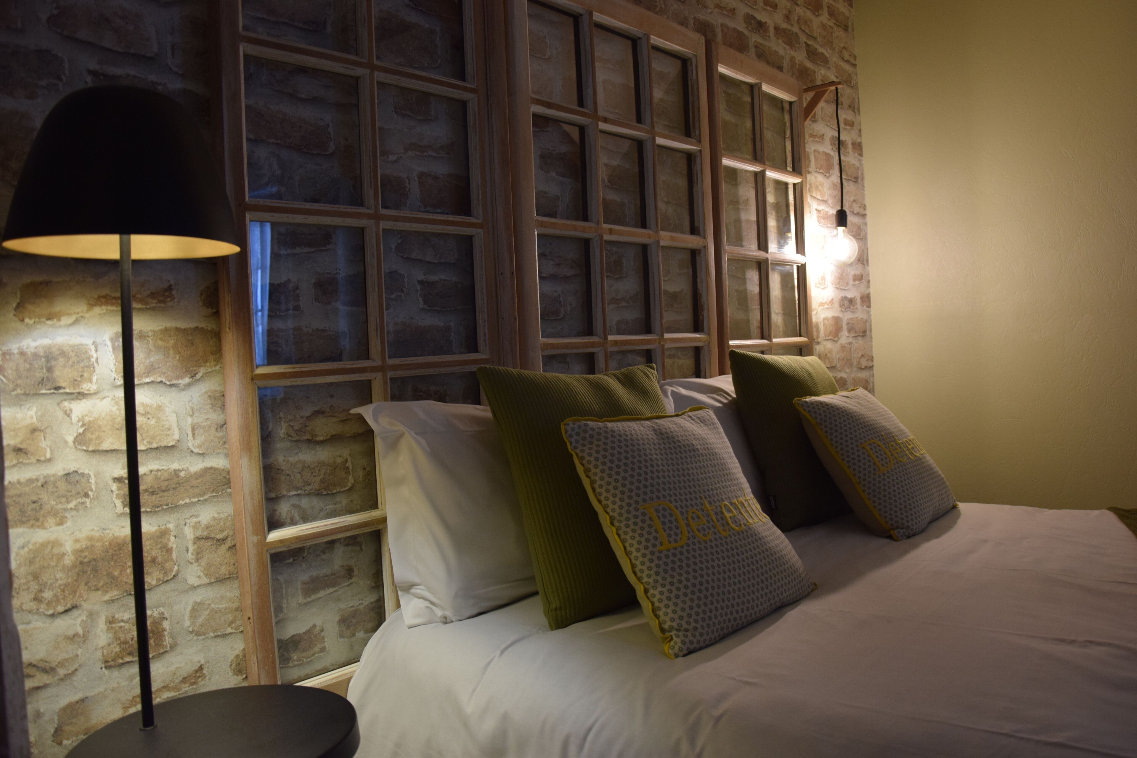 A bedroom with a brick wall, a large window with multiple panes, a floor lamp, and several pillows on the bed.