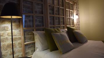 A bedroom with a brick wall, a large window with multiple panes, a floor lamp, and several pillows on the bed.