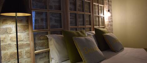 A bedroom with a brick wall, a large window with multiple panes, a floor lamp, and several pillows on the bed.