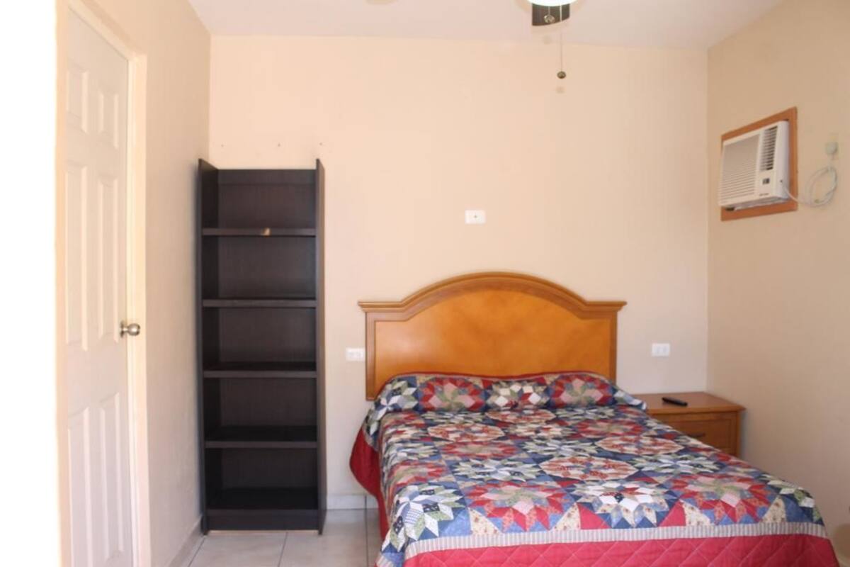 Un dormitorio con cama, cabecera de madera, mesita de noche, aire acondicionado de pared y estantería empotrada.