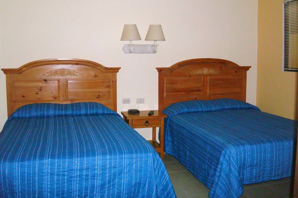 Una habitación con dos camas de madera, cada una con cubrecama azul, una mesita de noche con lámpara, y una lámpara de pared.