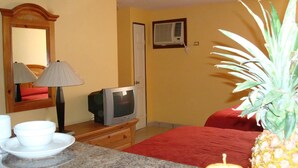 Een hotelkamer met een televisie, een spiegel, een rood sprei, een keukenhoek met een granieten aanrecht en een ananas op het aanrecht.