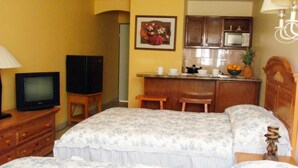 Een hotelkamer met twee bedden, een televisie, een bureau met stoel, een magnetron, en een kitchenette met een spoelbak en een schaal fruit.