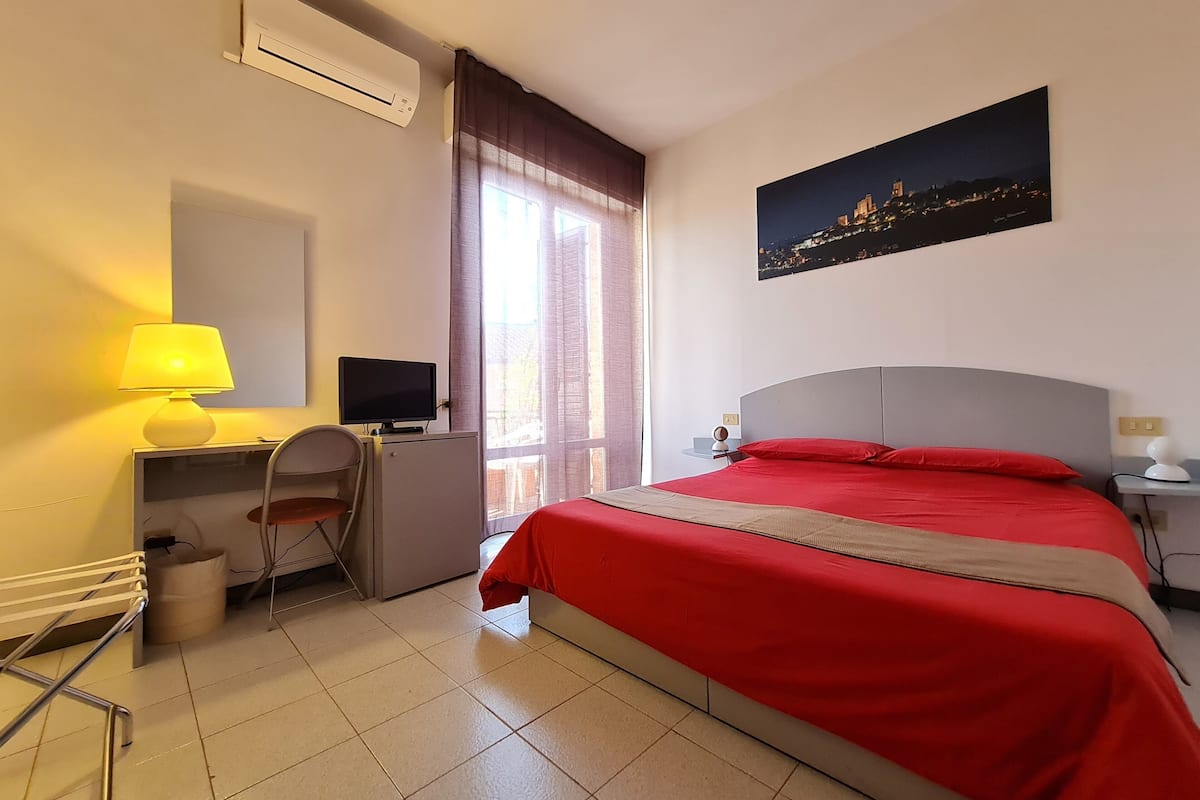 A hotel room with a bed, a desk with a computer, an air conditioner, and a window with curtains.