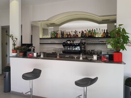Zona de bar moderna con techo curvo, una máquina de café y una variedad de botellas y vasos en estantes. Hay dos taburetes de bar negros y plantas en macetas.
