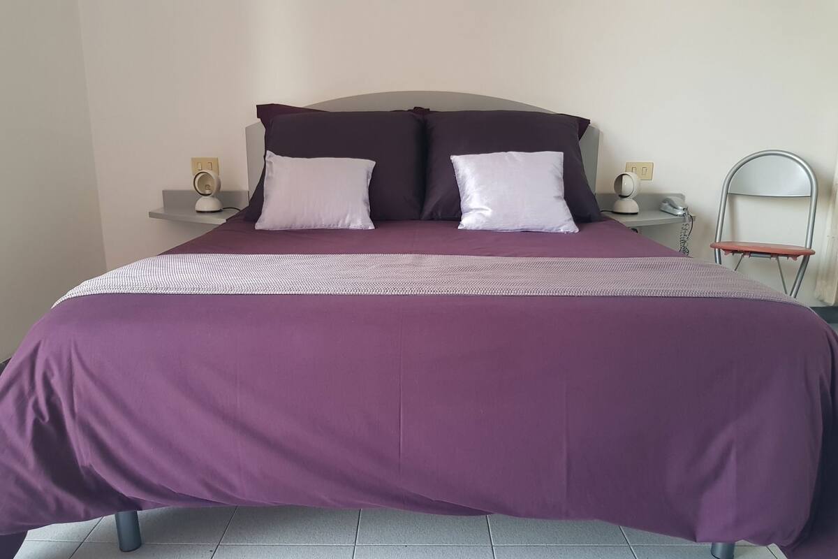 A bed with a purple bedspread and two white pillows, a bedside table with a lamp, and a chair.