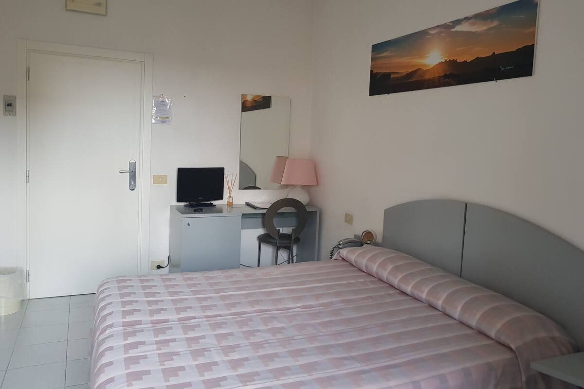 A bedroom with a bed, a desk with a computer, a chair, a lamp, and a painting of a sunset.