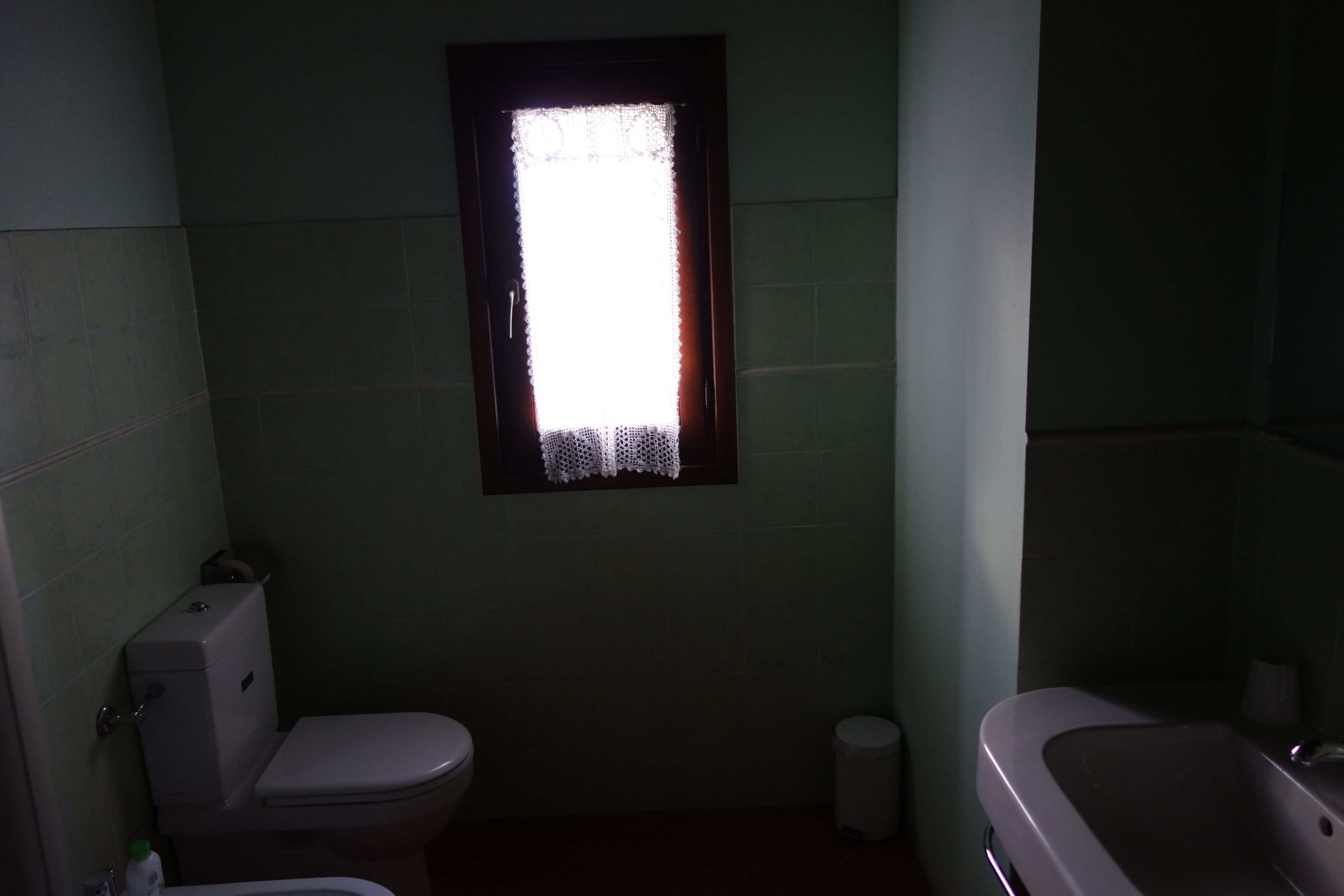 Un bagno piccolo e poco illuminato, con il water, un lavandino e una finestra con tenda.