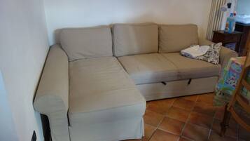 Un divano beige componibile con un poggiapiedi, un tavolino in legno con una lampada e una tovaglia colorata su una sedia.