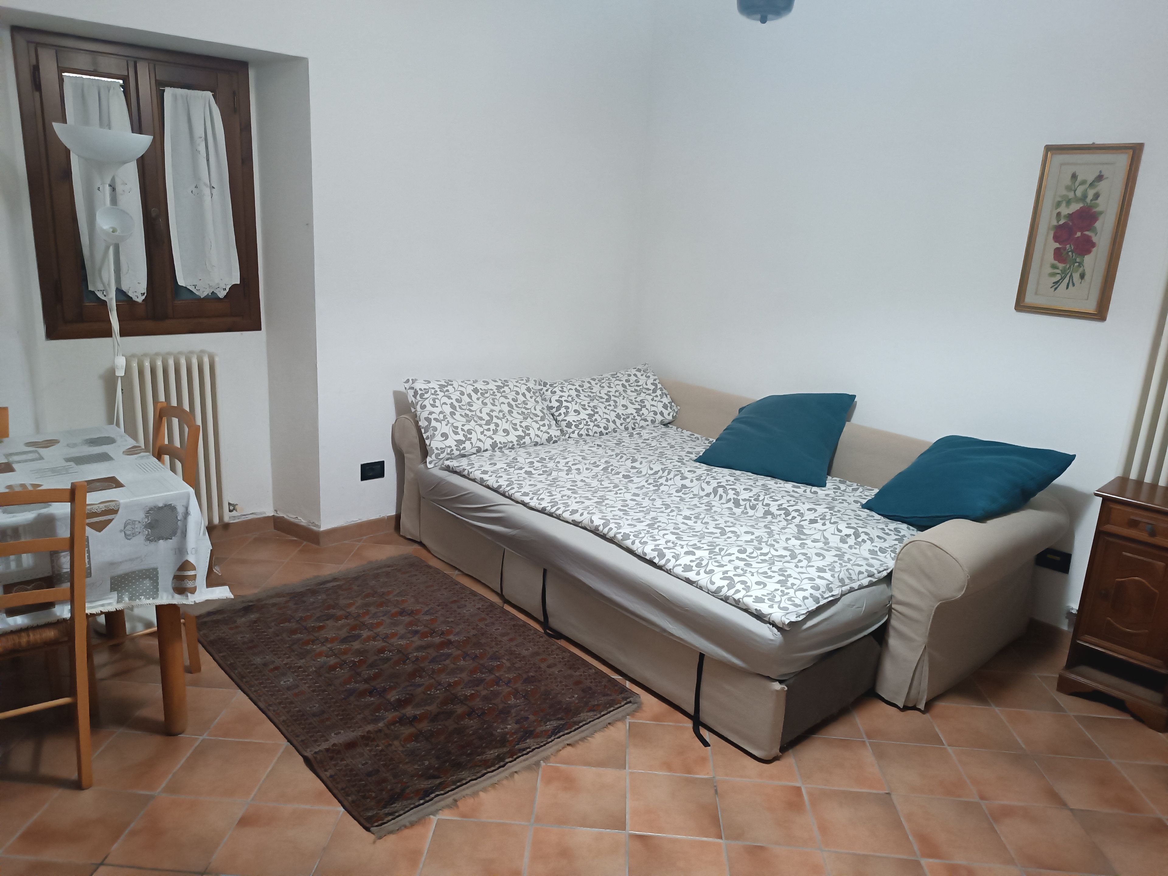Un apartamento pequeño de una sola habitación, con sofá cama, mesa de comedor con sillas, radiador y ventana con cortinas.