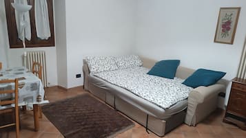 Un apartamento pequeño de una sola habitación, con sofá cama, mesa de comedor con sillas, radiador y ventana con cortinas.