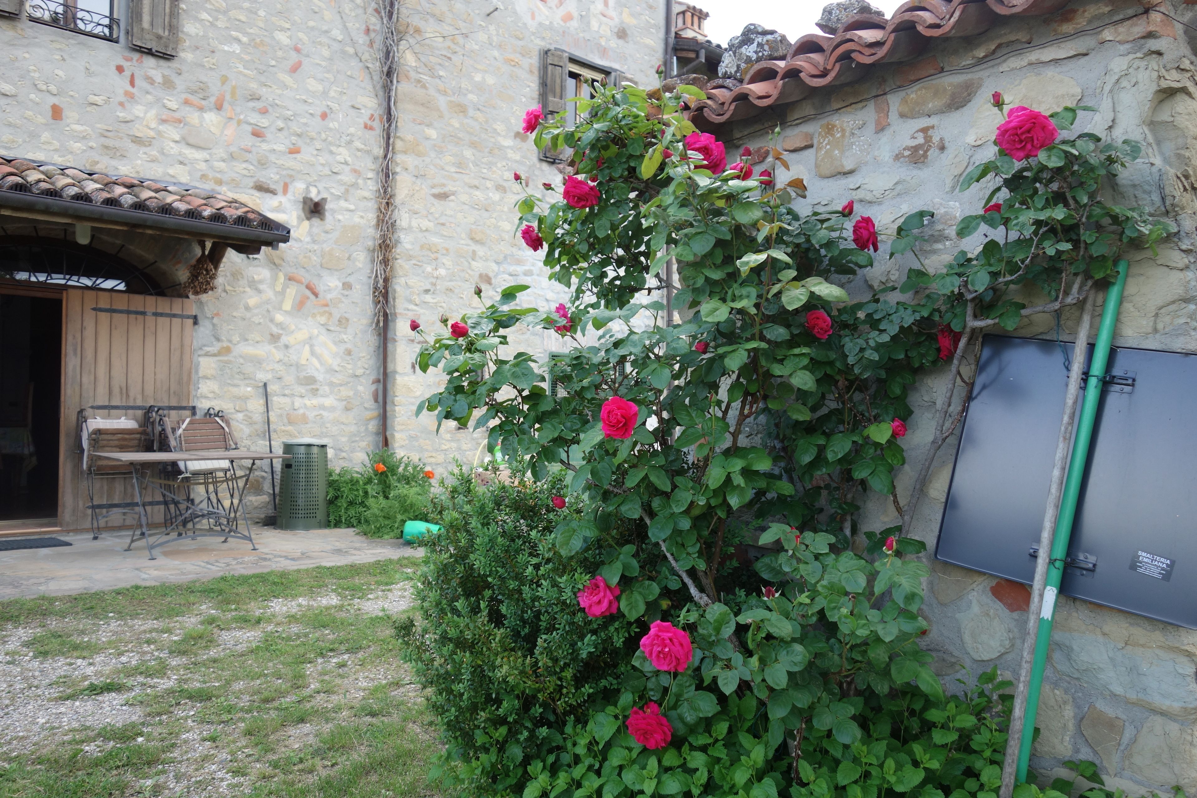 Un edificio de piedra con tejado de baldosas, una mesa y sillas de madera afuera, y un arbusto con flores rosadas.