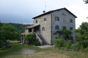 A stone building with a staircase leading to a porch, surrounded by greenery and hills in the background. - Bed and breakfast Monte Bibele (Monterenzio)