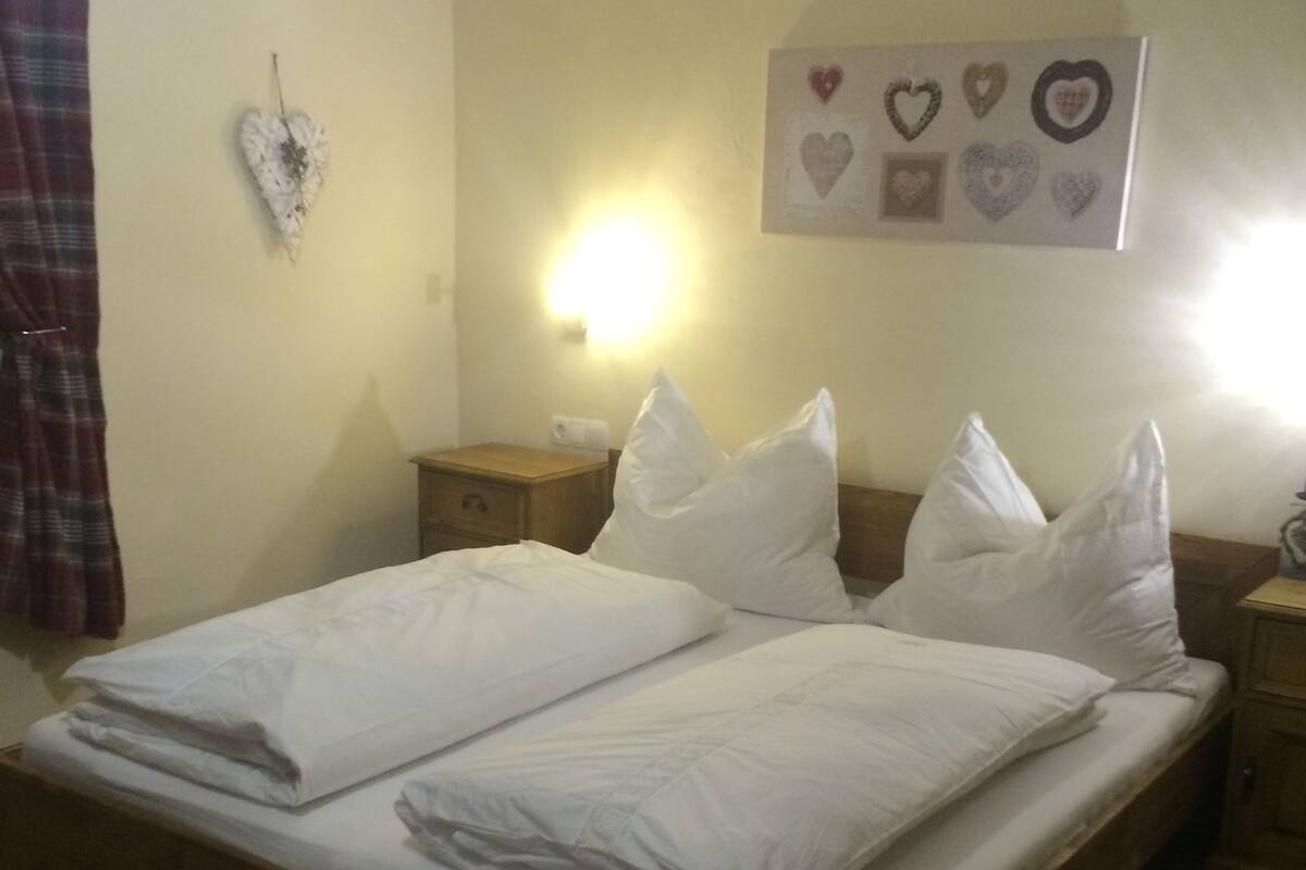 Una cama doble con ropa de cama blanca, una mesita de noche con lámpara, una decoración con forma de corazón en la pared y una cortina a cuadros.