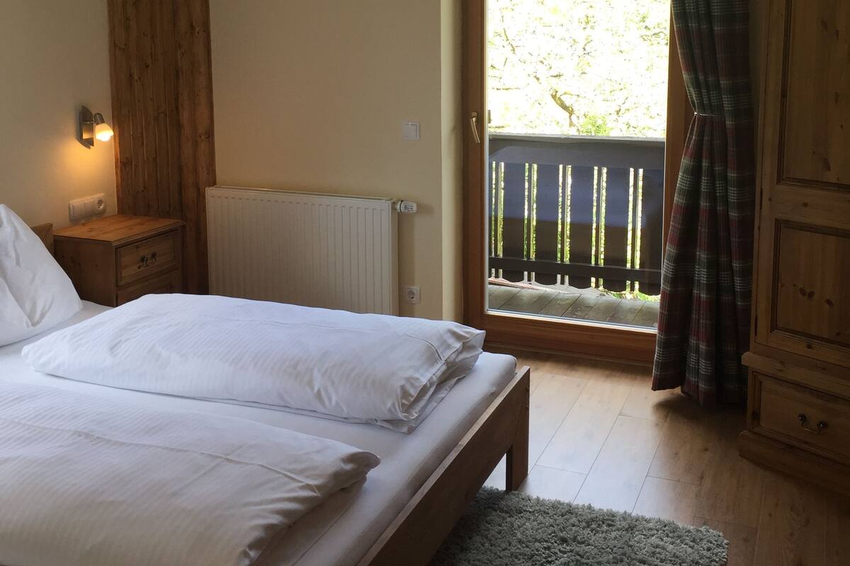 Un dormitorio con una cama doble, una mesita de noche de madera, un radiador, una ventana que da a un balcón y una puerta con cortina escocesa.