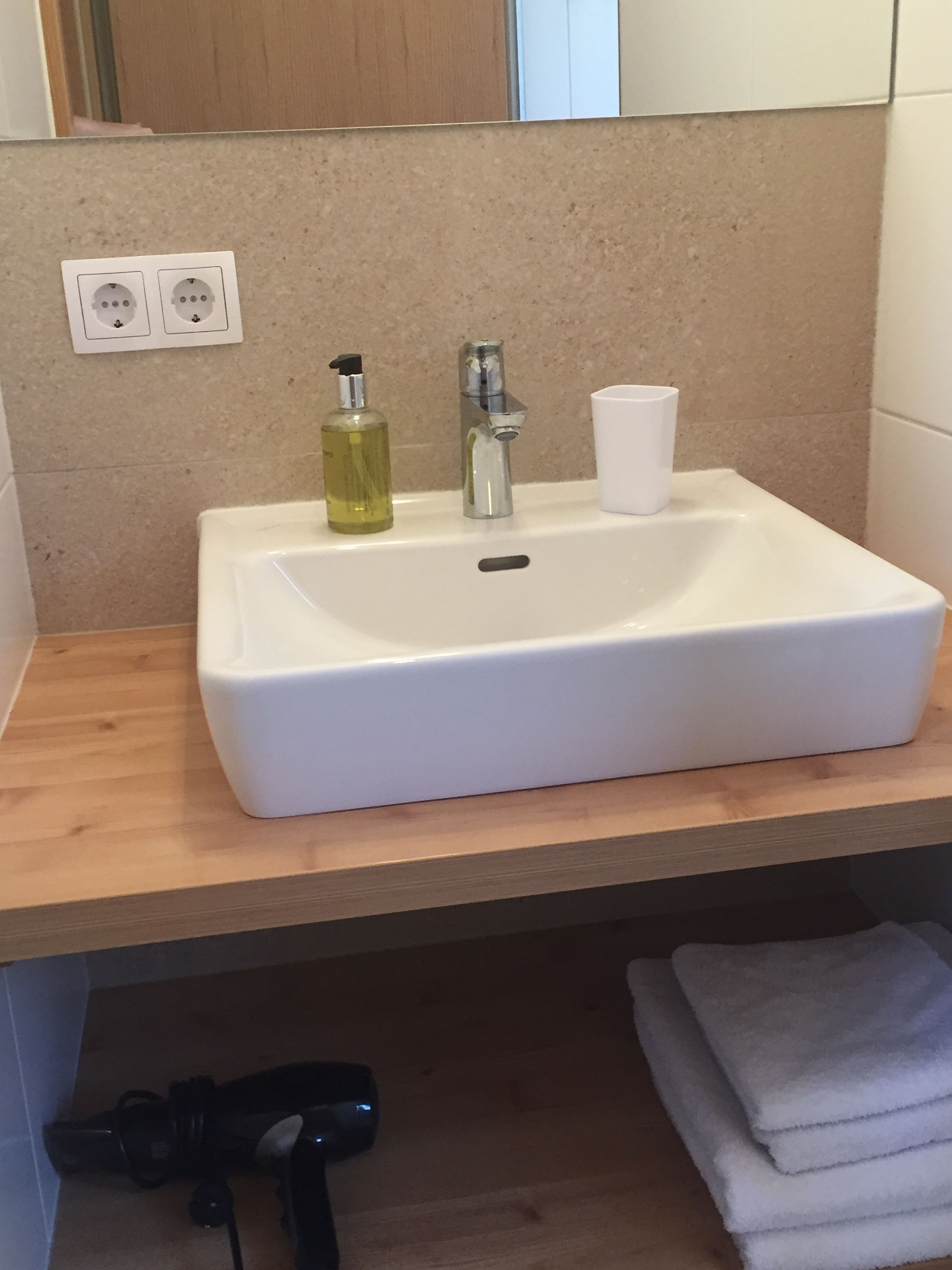 Un bany amb un lavabo blanc, una ampolla de sabó líquid i una tassa. Hi ha una prestatgeria de fusta sota el lavabo, i una tovallola apilada a la dreta.