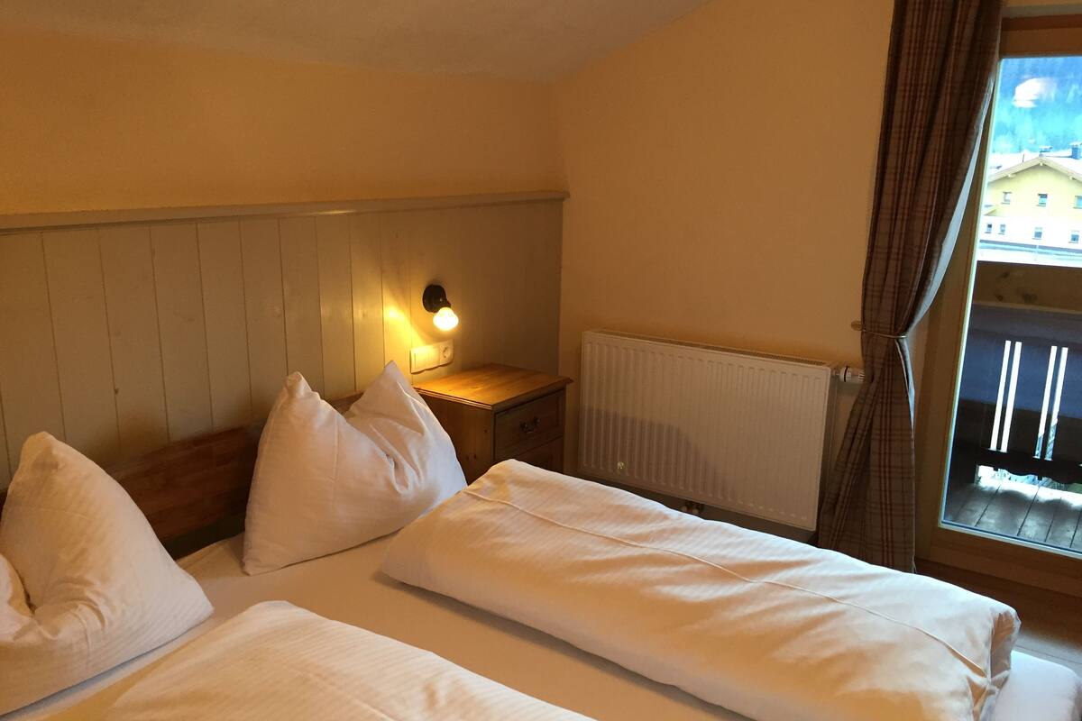 Habitación de hotel con dos camas, una mesita de noche con lámpara, un radiador y una ventana con cortinas.