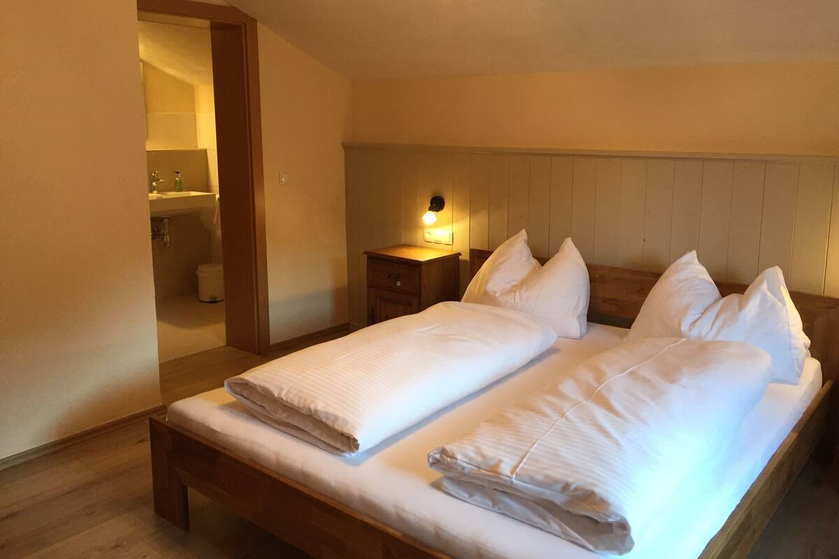 Un dormitorio con cama de madera, ropa de cama blanca, una mesita de noche con lámpara y vistas al baño.
