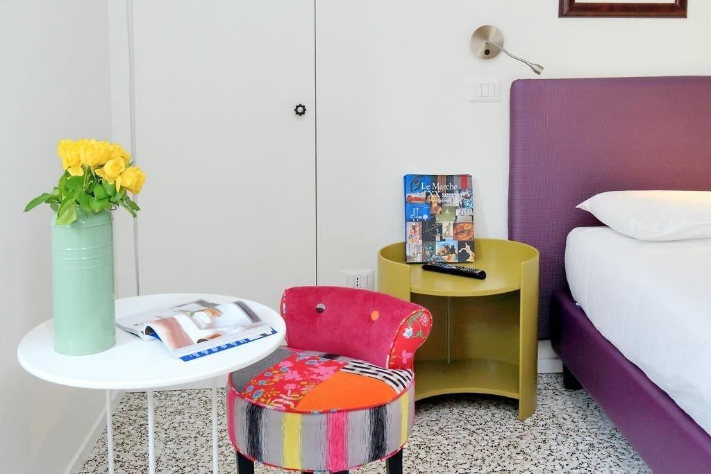 Quarto de hotel com cama, uma mesinha com uma revista, uma cadeira, um vaso com flores e um livro em uma mesinha de cabeceira.