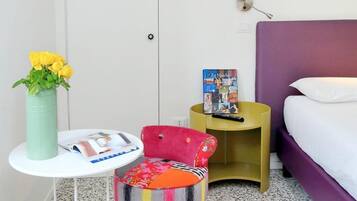 Quarto de hotel com cama, uma mesinha com uma revista, uma cadeira, um vaso com flores e um livro em uma mesinha de cabeceira.