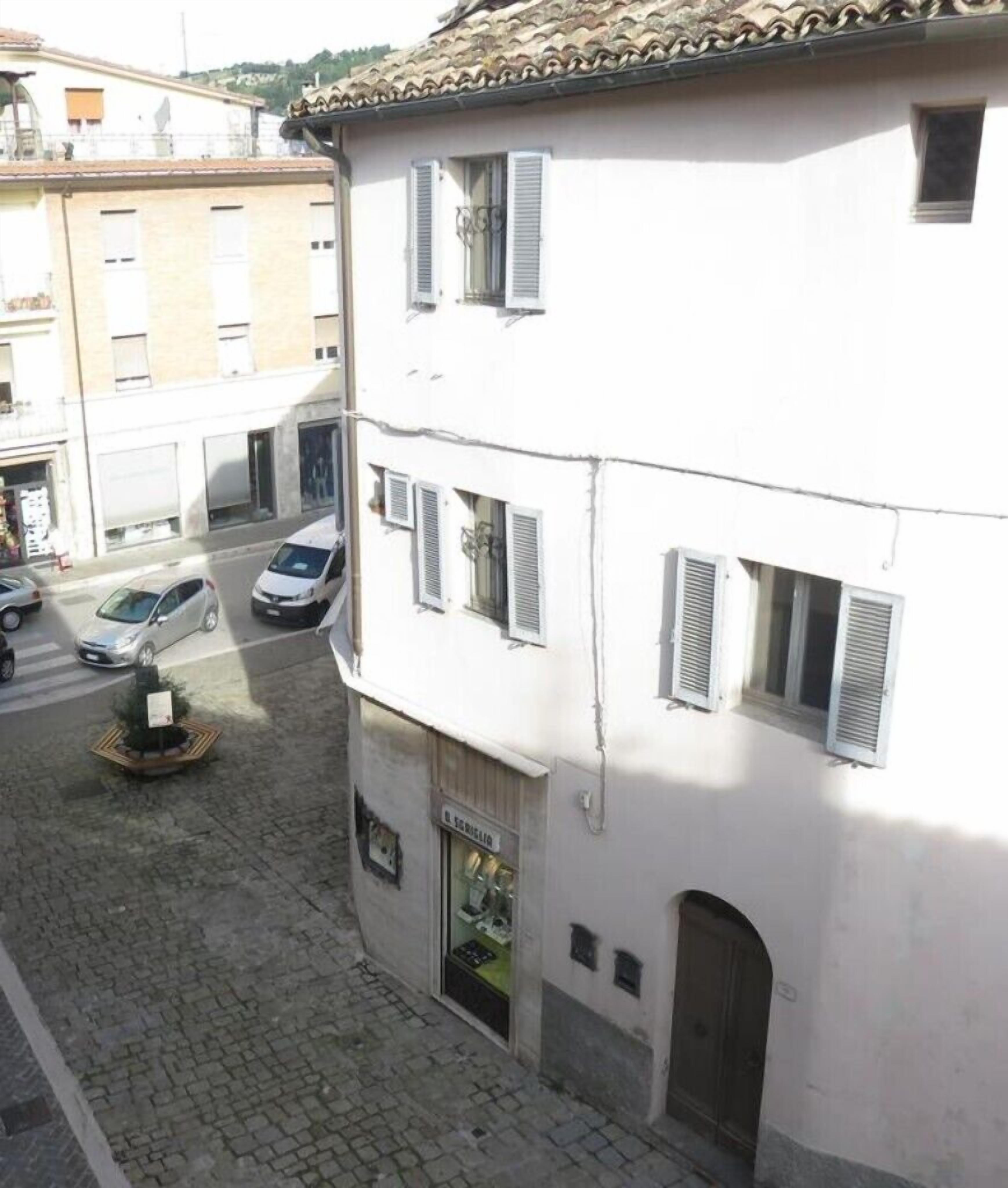 A narrow street with a white building featuring multiple windows with shutters, a visible storefront, and a cobblestone pavement.