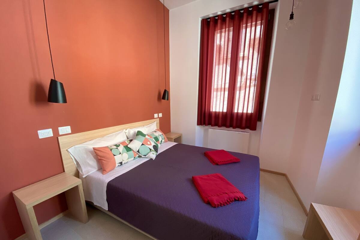 Un dormitorio con cama, mesitas de noche, una ventana con cortinas y una lámpara colgante.