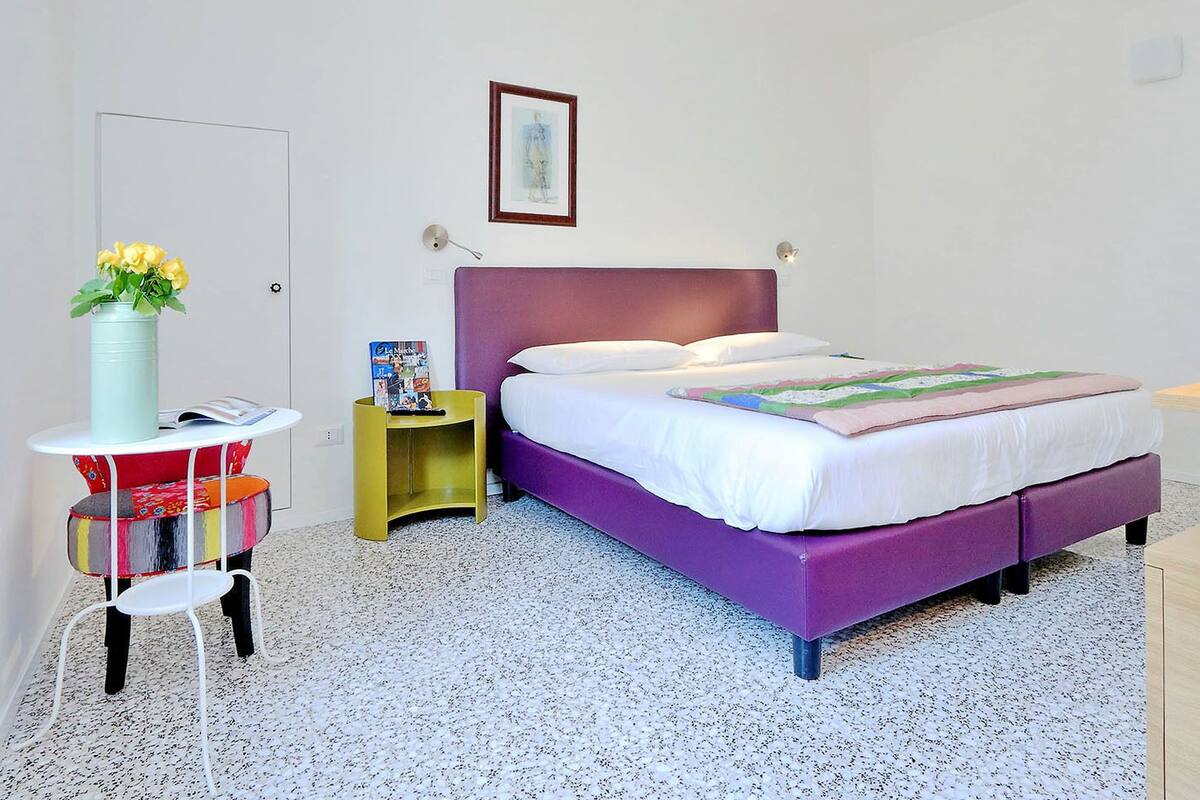 Un dormitorio con cama morada, ropa de cama blanca, una mesita con un florero y un cuadro en la pared.