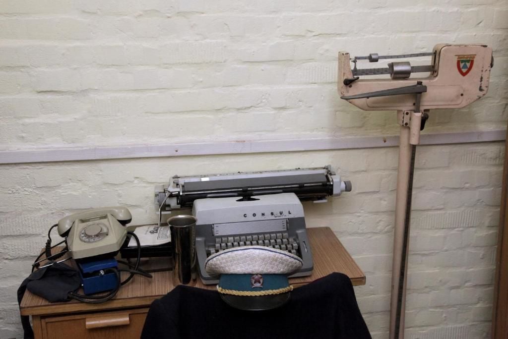 A vintage typewriter, a rotary phone, and a small tool on a wooden desk against a white brick wall.