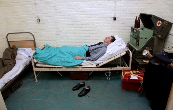 A single-bed room with a metal frame, a teal blanket, a pair of shoes, a red toolbox, and a medical device on a table.