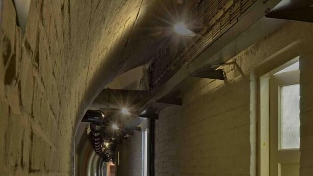 A narrow, dimly lit hallway with brick walls and a wooden floor, leading to a door at the end.