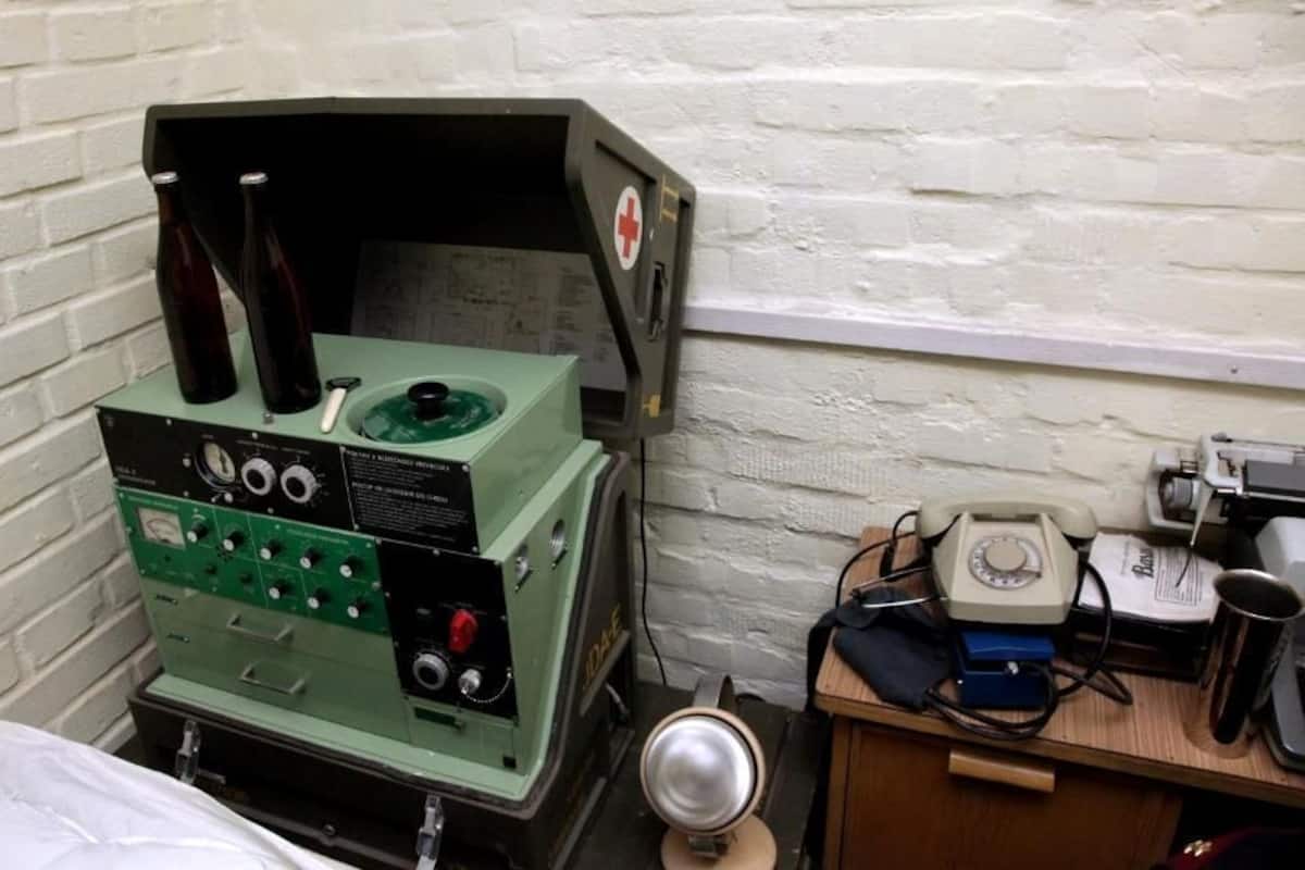 A room with a bed, a green machine with dials and buttons, a telephone, and a small table with a cup and papers.