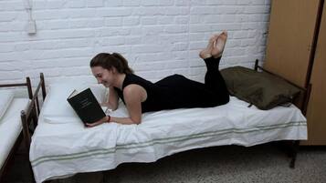 Una persona recostada en una cama con un libro, en una habitación con pared de ladrillo blanco y puerta de madera.