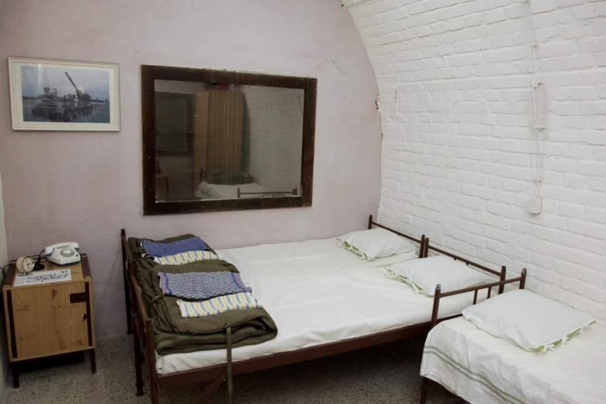 A small, single-room with a bunk bed, a wooden nightstand with a telephone, a framed picture on the wall, and a window with a curtain.