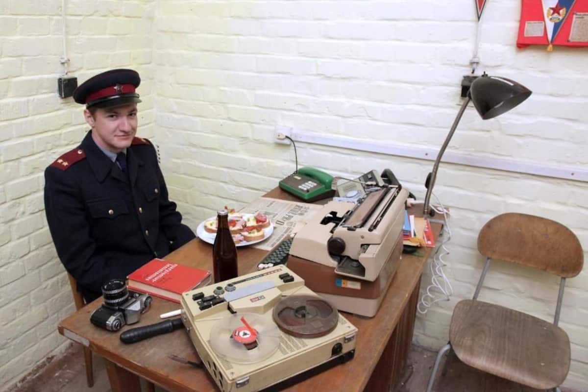 A person in uniform sitting at a desk with a typewriter, a camera, and a bottle.